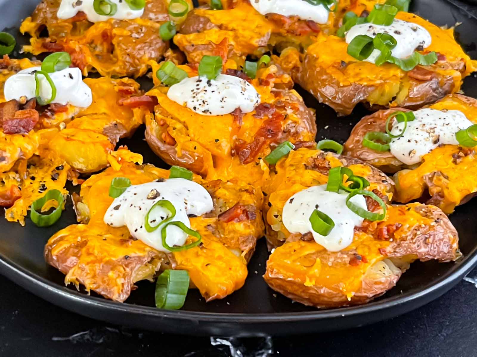 Loaded smashed potatoes on a dark platter garnished with diced green onions and sour cream.