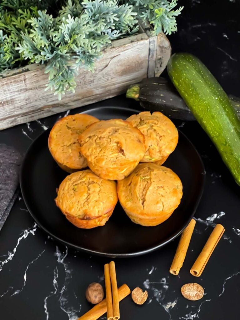 Zucchini muffins on a dark plate with some cinnamon sticks and nutmeg.