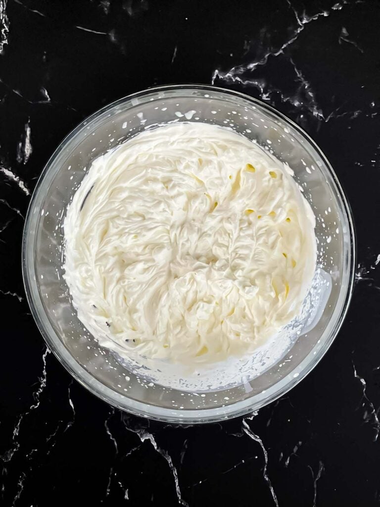 Heavy cream whipped to stiff peaks in a glass mixing bowl on a dark surface.