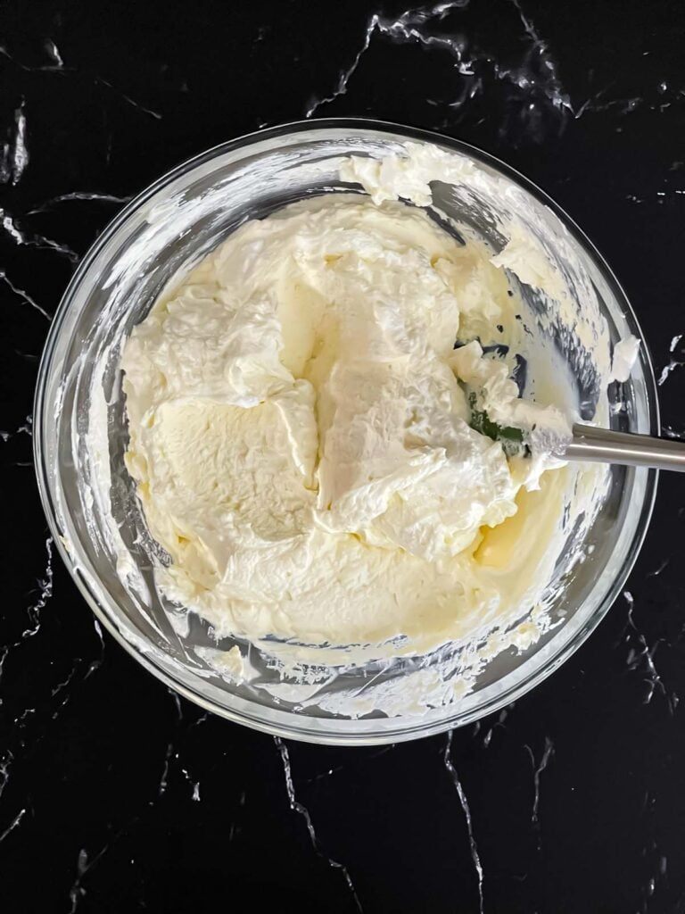 Lemon curd folded into the whipped cream in a glass mixing bowl on a dark surface.
