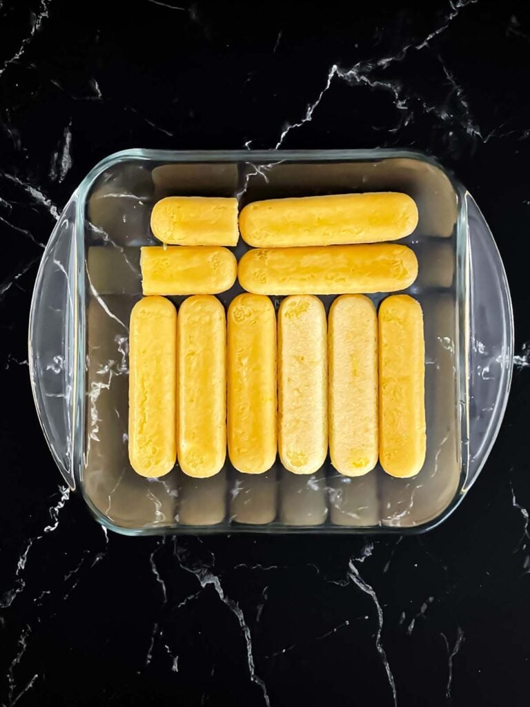 Dipped lady fingers lining a glass baking dish on a dark surface.