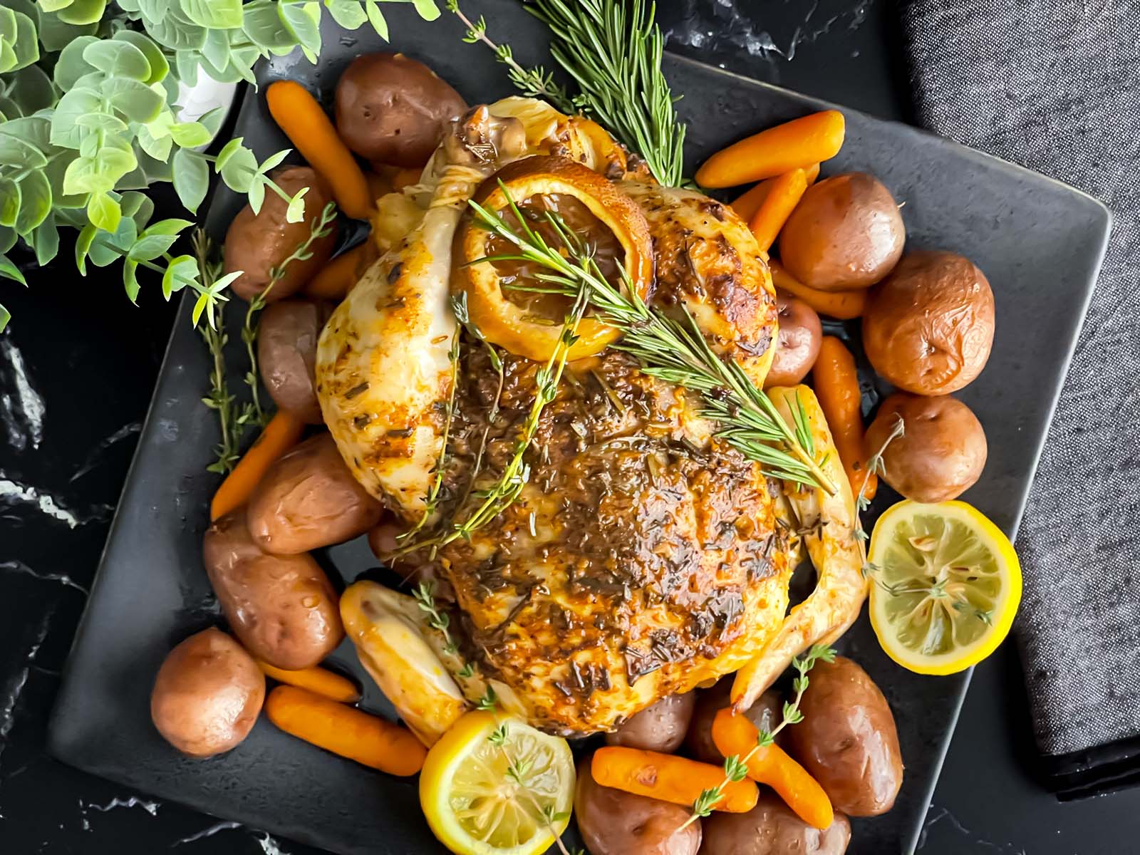 Whole lemon and herb roasted chicken on a dark plate surrounded by potatoes and carrots.