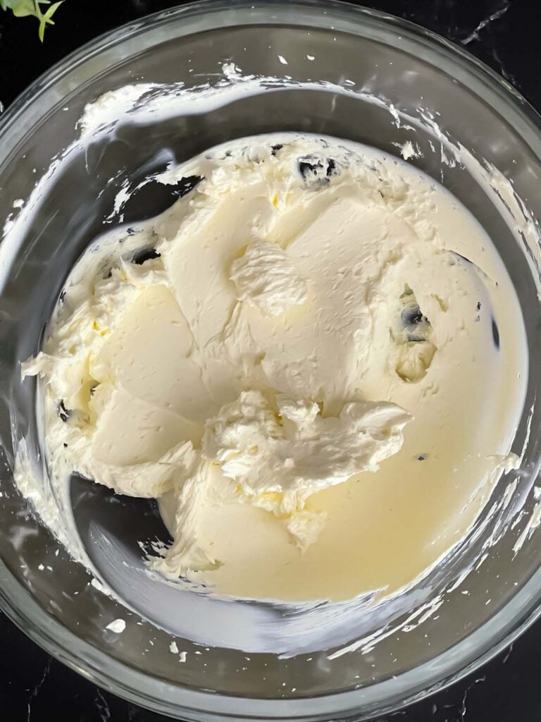 Softened cream cheese for the vegetable tortilla roll ups recipe beaten smooth in a large glass bowl on a dark surface.