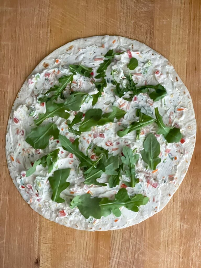 One tortilla on a wooden cutting board layered with the cream cheese mixture and baby arugula.