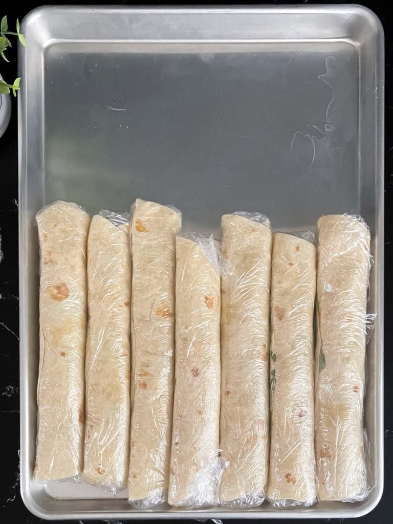 Veggie tortilla roll ups wrapped individually in plastic wrap, lined up on a baking sheet.