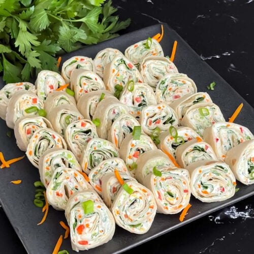 Veggie tortilla roll ups lined up on a square dark plate garnished with diced green onions and shredded carrots.