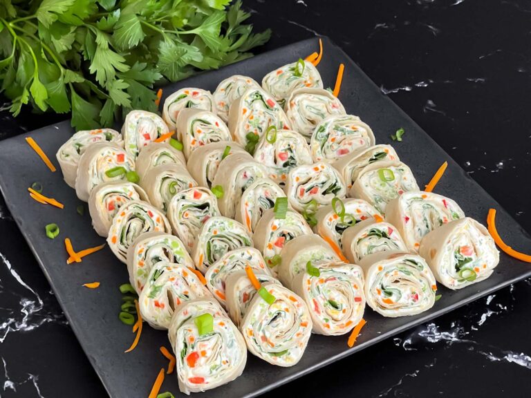 Veggie tortilla roll ups lined up on a square dark plate garnished with diced green onions and shredded carrots.