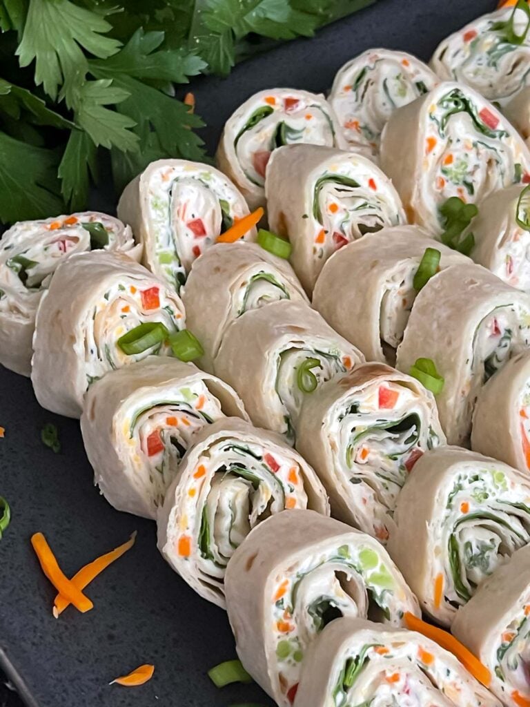 Veggie tortilla roll ups lined up on a square dark plate garnished with diced green onions and shredded carrots.