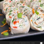 Veggie tortilla roll ups lined up on a square dark plate garnished with diced green onions and shredded carrots.