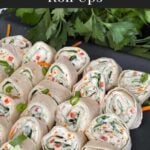 Veggie tortilla roll ups lined up on a square dark plate garnished with diced green onions and shredded carrots.