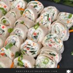 Veggie tortilla roll ups lined up on a square dark plate garnished with diced green onions and shredded carrots.