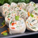 Veggie tortilla roll ups lined up on a square dark plate garnished with diced green onions and shredded carrots.