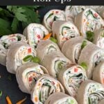 Veggie tortilla roll ups lined up on a square dark plate garnished with diced green onions and shredded carrots.