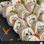 Veggie tortilla roll ups lined up on a square dark plate garnished with diced green onions and shredded carrots.
