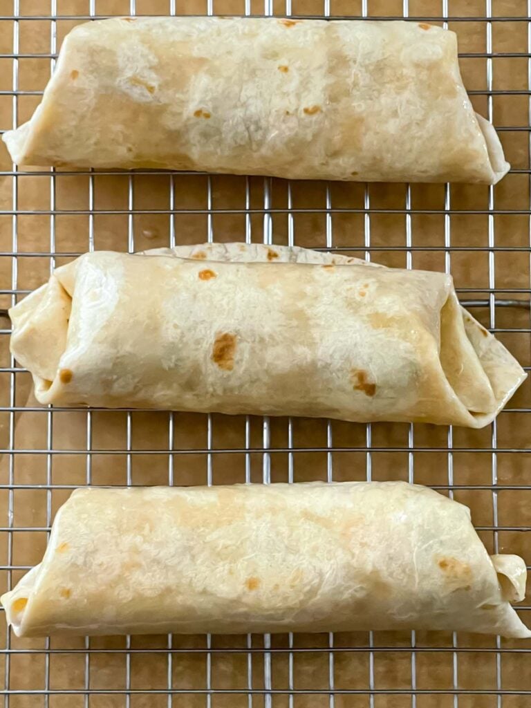Three chicken chimichangas on a wire rack over a parchment paper lined baking sheet.