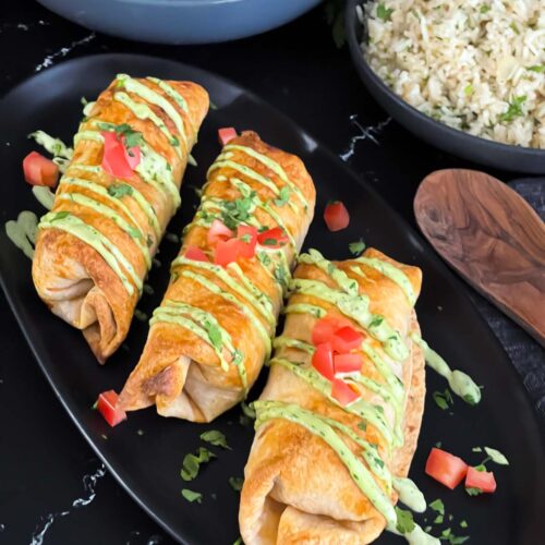 Baked chicken chimichangas on a dark oval platter garnished with avocado crema, diced tomatoes and cilantro.