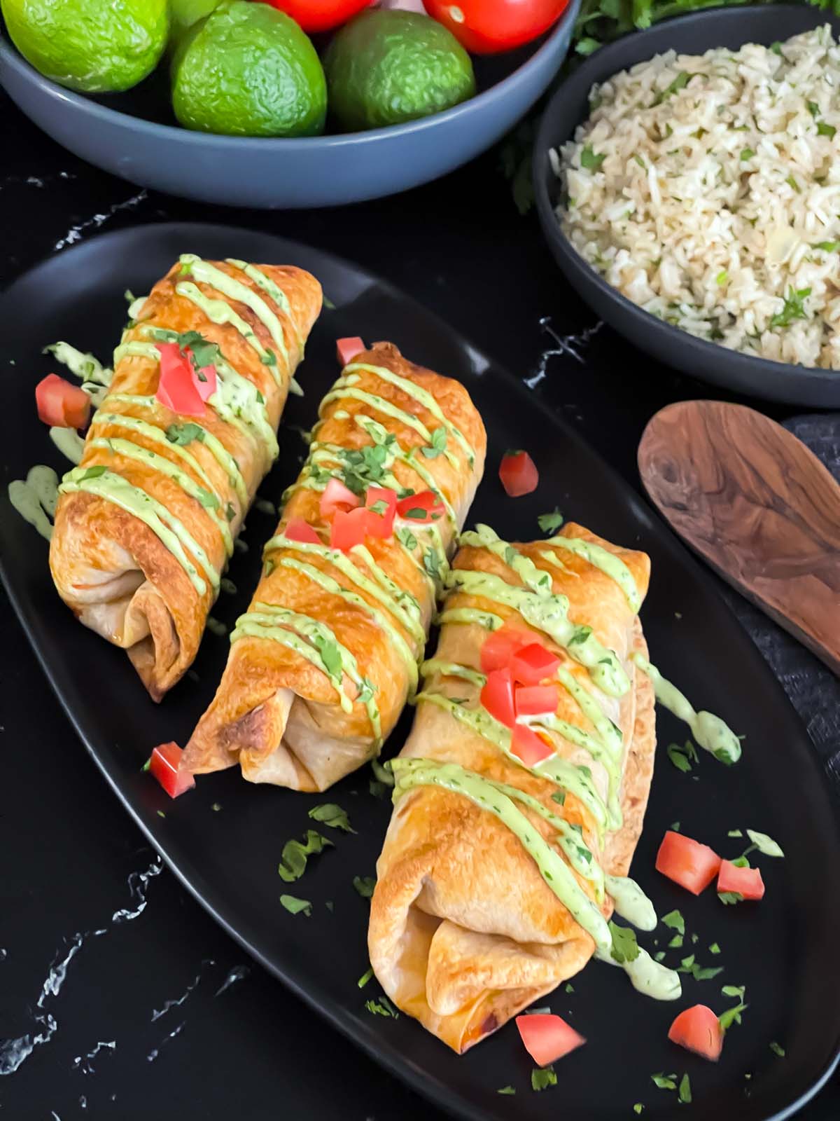 Baked chicken chimichangas on a dark oval platter garnished with avocado crema, diced tomatoes and cilantro.