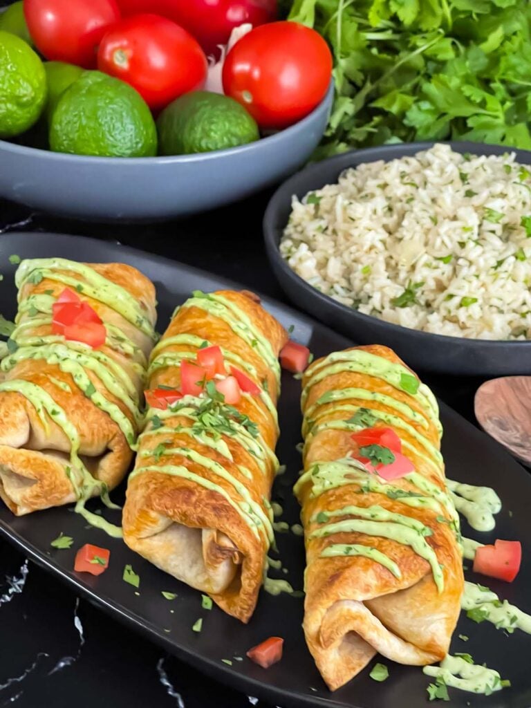 Baked chicken chimichangas on a dark oval platter garnished with avocado crema, diced tomatoes and cilantro.