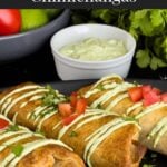 Baked chicken chimichangas on a dark oval platter garnished with avocado crema, diced tomatoes and cilantro.