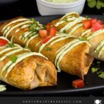 Baked chicken chimichangas on a dark oval platter garnished with avocado crema, diced tomatoes and cilantro.