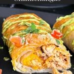Baked chicken chimichangas on a dark oval platter garnished with avocado crema, diced tomatoes and cilantro.
