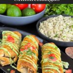 Baked chicken chimichangas on a dark oval platter garnished with avocado crema, diced tomatoes and cilantro.