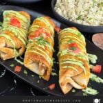 Baked chicken chimichangas on a dark oval platter garnished with avocado crema, diced tomatoes and cilantro.