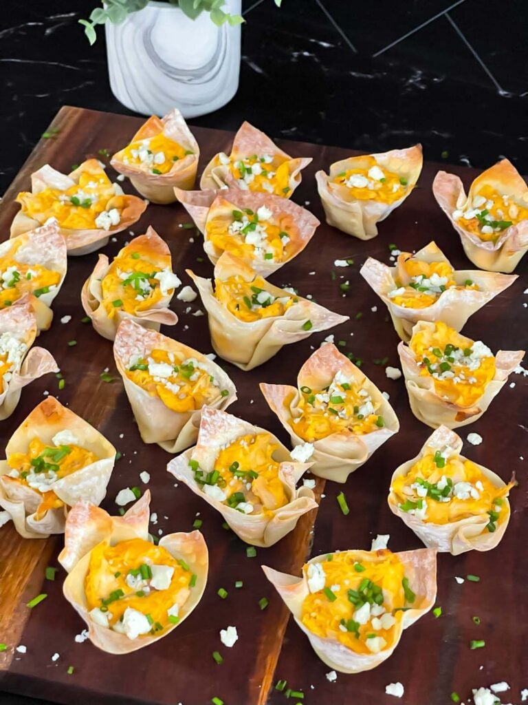 Buffalo chicken wontons garnished with chives and blue cheese crumbles on a cutting board.