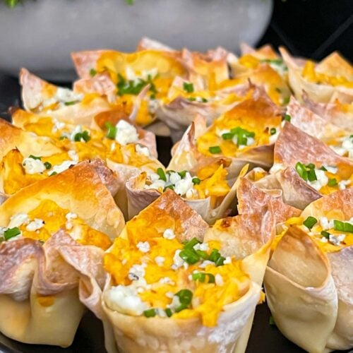 Buffalo chicken wontons garnished with chives and blue cheese crumbles on a cutting board.