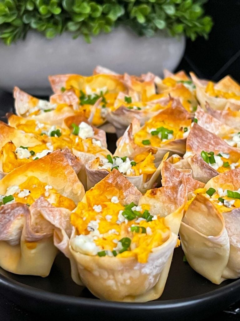 Buffalo chicken wontons garnished with chives and blue cheese crumbles on a cutting board.