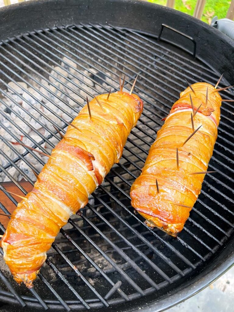 Bacon wrapped pork tenderloin cooking over indirect heat on a grill.