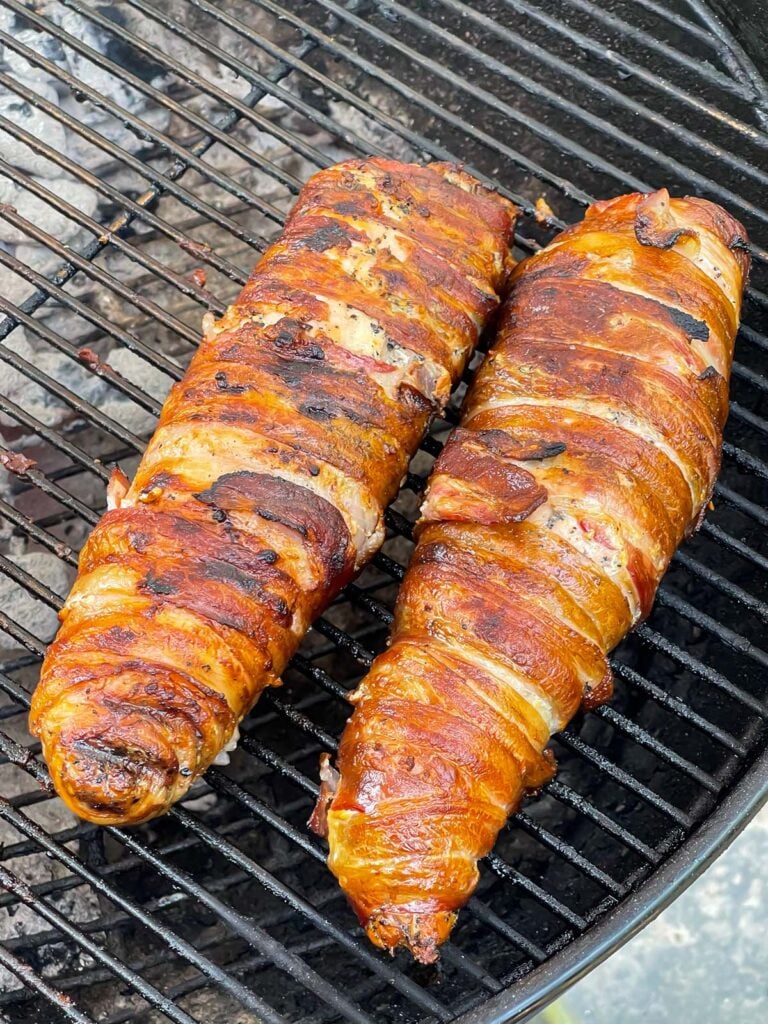 Bacon wrapped pork tenderloin cooking over indirect heat on a grill.
