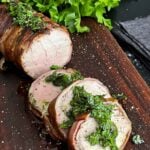 Bacon wrapped pork tenderloin with a chimichurri on a dark cutting board.