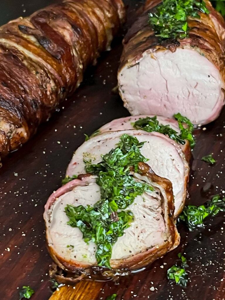 Bacon wrapped pork tenderloin with a chimichurri on a dark cutting board.