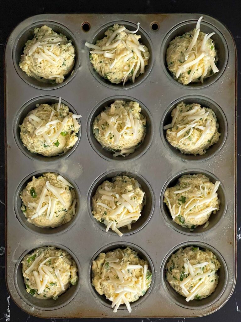 Herb and cheese muffin batter divided into 12 muffin cups topped with shredded fontina cheese.
