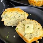 Herb and cheese muffin cut in half topped with butter on a dark plate.