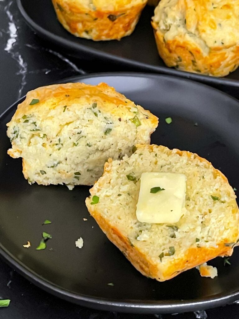 Herb and cheese muffin cut in half topped with butter on a dark plate.