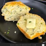 Herb and cheese muffin cut in half topped with butter on a dark plate.