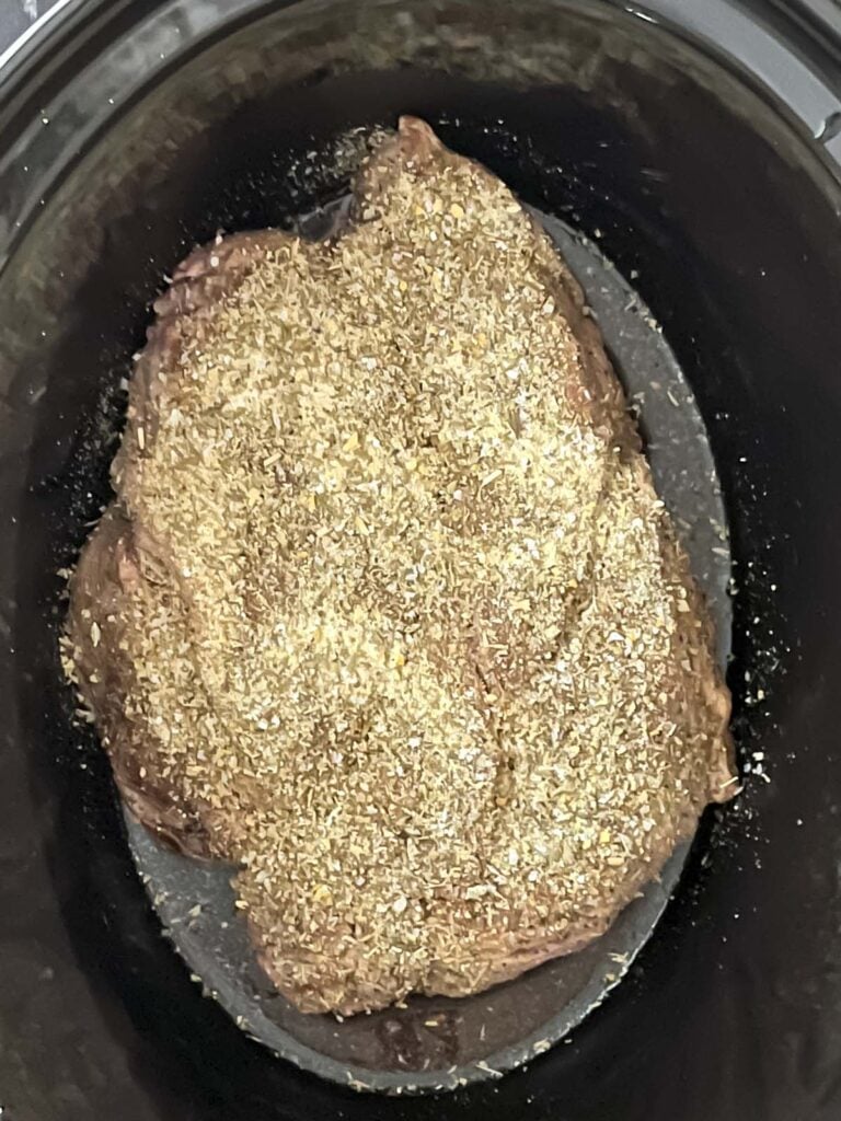 Chuck roast sprinkled with seasonings in the crock of a slow cooker.