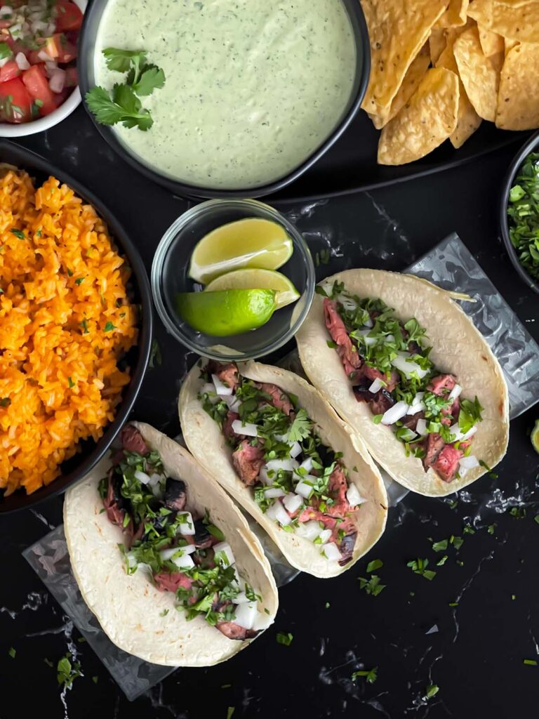 Flank steak street tacos with rice, chips, and garnish on a dark background.