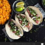 Flank steak street tacos with rice, chips, and garnish on a dark background.