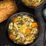 Chicken noodle soup in a dark bowl on a dark surface.