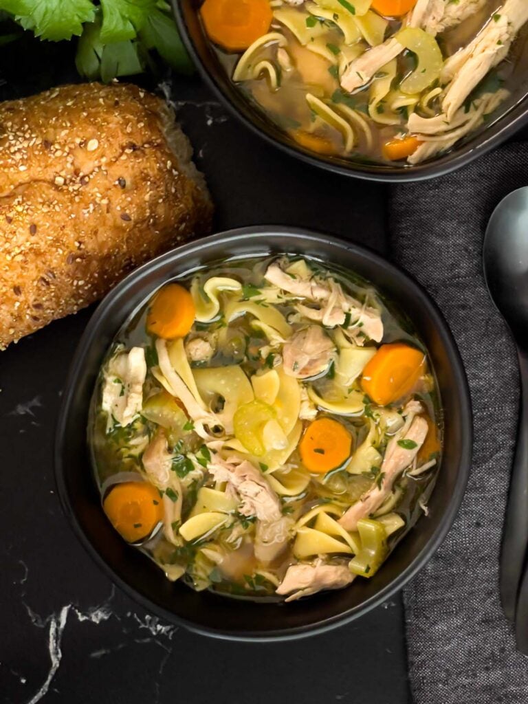 Chicken noodle soup in a dark bowl on a dark surface.