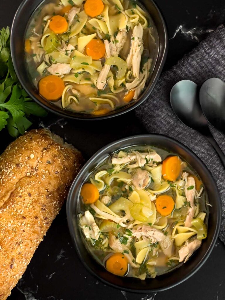 Chicken noodle soup in a dark bowls on a dark surface.