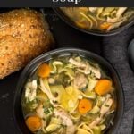 Chicken noodle soup in a dark bowl on a dark surface.