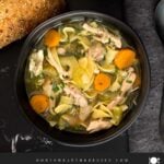 Chicken noodle soup in a dark bowl on a dark surface.