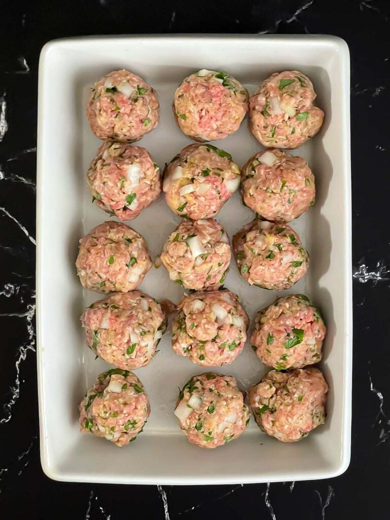 Unbaked parmesan meatballs in a light casserole dish on a dark surface.