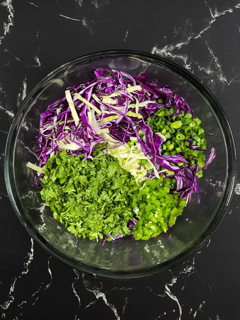 Slaw ingredients in a glass bowl before mixing, including cabbage, cilantro, green onion, and jalapeño.