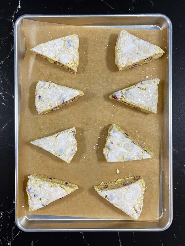 Unbaked cranberry orange scones brushed with heavy cream on a parchment paper lined baking sheet.