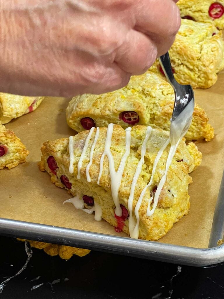 Orange glaze being drizzled over a cranberry orange scone on the prepared baking sheet.